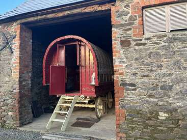 Une roulotte rouge traditionnelle et ornée, au toit incurvé, se dresse dans une grange en pierre, avec des marches en bois menant à ses portes ouvertes.