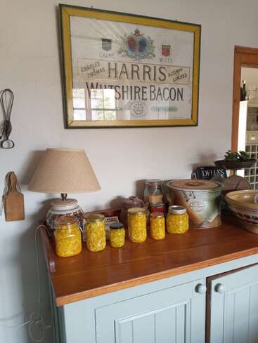 Eine gemütliche Küchenszene zeigt eine Holztheke mit Gläsern voller eingelegter gelber Gemüsesorten, eine Lampe, Retro-Küchenutensilien und darüber ein Vintage-Schild mit der Aufschrift „Harris Wiltshire Bacon“.