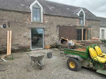 Ein altes Steinhaus, das gerade renoviert wird, mit Baugeräten, einem John Deere Gator, einer Schubkarre, einem Betonmischer, Holzbrettern und Kies, der den Boden bedeckt.