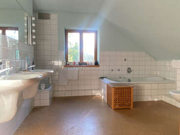 Este banheiro espaçoso tem pias duplas, uma banheira com azulejos brancos, uma janela de madeira, luz natural, um cesto de vime, espelhos e um design clean e moderno.