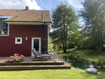 A charming red house with a yellow-tiled roof, wooden deck, lounge chairs, blooming flowers, and a small pond, surrounded by lush green trees and grass.