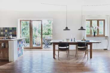 This image shows a bright, modern kitchen and dining area with wooden floors, large windows, a wooden table, colorful tiles, pendant lights, and outdoor garden views.