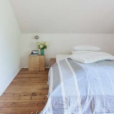 L'image représente une chambre à coucher sereine avec un lit, une table de nuit et un plancher en bois, avec une couverture à rayures bleues et blanches, des oreillers et des draps d'un blanc immaculé.