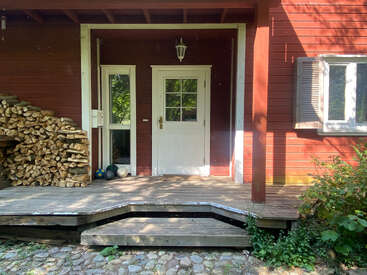 A rustic wooden porch with a stacked firewood pile, white front door, lantern, sports balls, red siding, cobblestone path, greenery, and sunlight casting shadows.