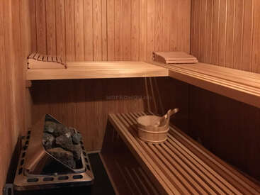 This image shows the interior of a wooden sauna. There are wooden benches, a bucket with a ladle, and a heater with stones for steam.