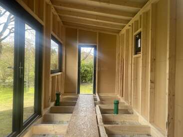 This image shows the inside of a small wooden structure under construction, featuring exposed framing, open windows, a door, and green pipes on the unfinished floor.