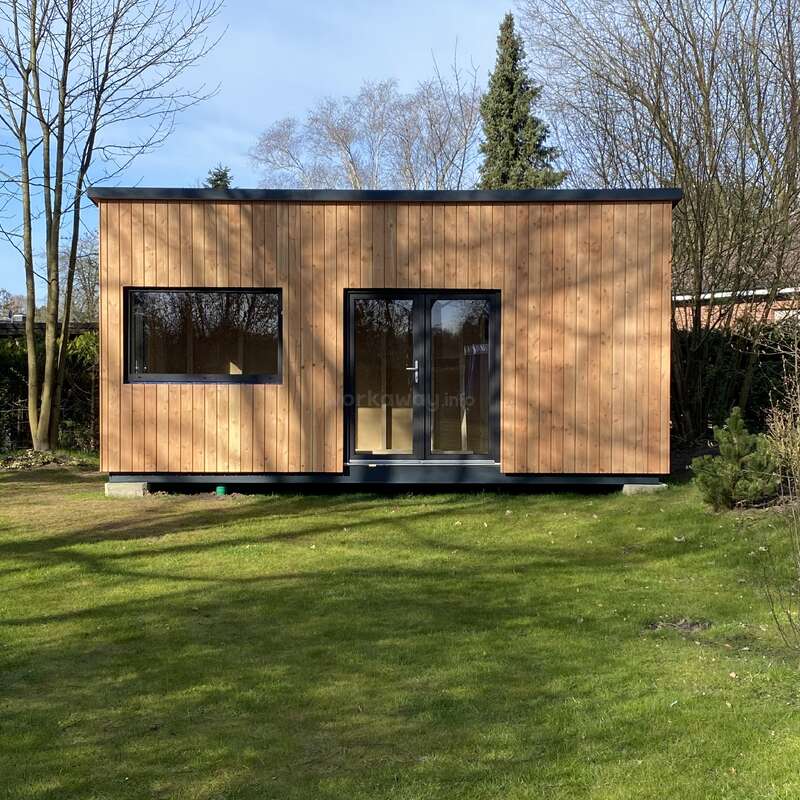 A modern, small wooden cabin with large windows stands on a grassy lawn, surrounded by leafless trees and shrubs, under a clear blue sky.