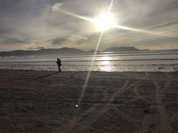 A imagem retrata uma cena serena na praia com uma pessoa passeando ao longo da costa, tendo como pano de fundo montanhas e um sol radiante brilhando intensamente no céu.