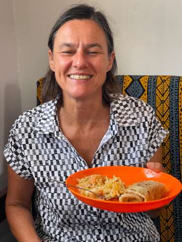 Una mujer sonriente está sentada en el interior, sosteniendo alegremente un plato naranja con chapati y ensalada de verduras. Lleva una camisa con estampado en blanco y negro y parece disfrutar de su comida.