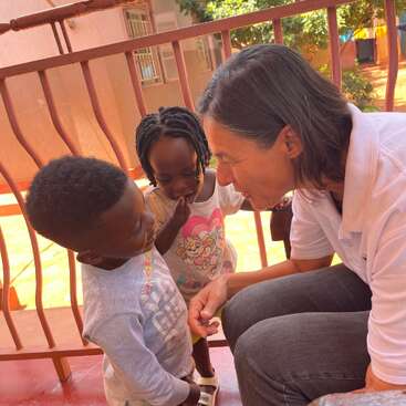 Eine Frau kniet sanft nieder, lächelt und geht herzlich auf zwei kleine Kinder zu. Die Szene spielt draußen, ist voller Sonnenlicht und strahlt Freundlichkeit, Zuneigung und Verbundenheit aus.