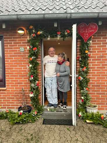 Una pareja posa sonriente en un portal decorado con flores y guirnaldas, celebrando su 25.º aniversario, tal y como indica el cartel rojo con forma de corazón que hay encima.