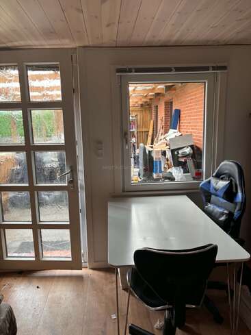 Una pequeña habitación con techo de madera, puerta blanca y ventana. Dentro hay una mesa blanca y dos sillas, y fuera hay un trastero abarrotado.