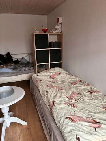 Una acogedora habitación con dos camas, una de ellas con ropa de cama de unicornios. Hay una mesa redonda blanca, suelo de madera, estanterías cúbicas y juguetes encima de la estantería.