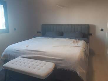 Une chambre à coucher moderne comprend un grand lit avec une tête de lit capitonnée grise, deux oreillers foncés, une couette blanche et un banc touffeté au pied.