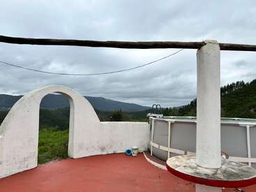 Esta imagen muestra una terraza exterior circular con paredes blancas, arco y columna alta. Hay una piscina redonda, vistas a la montaña, cielo nublado y suelo de baldosas rojas.