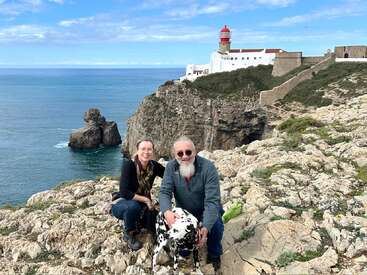 Una pareja sonriente con un perro dálmata posa en unos acantilados rocosos con vistas al océano, con un faro blanco en lo alto de la dramática costa bajo un cielo azul.