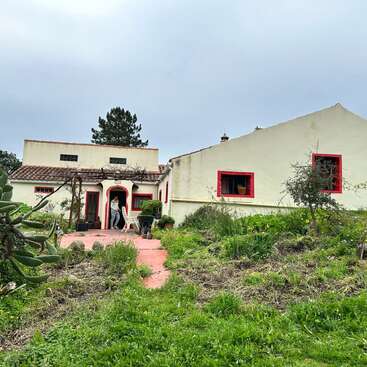 La imagen muestra una gran casa de color blanco roto con ribetes rojos y tejado de tejas, con un patio y un frondoso jardín en primer plano.