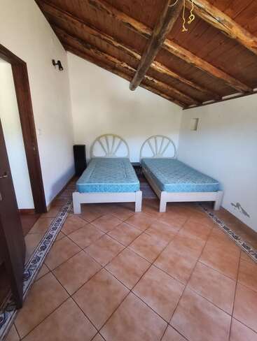 Cette image montre une chambre mansardée simple avec deux lits simples, un plafond incliné en bois, un sol carrelé, des murs blancs et une décoration minimale. La pièce est confortable et rustique.