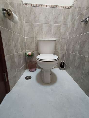 Cette image montre une petite salle de bains propre avec des murs carrelés de couleur beige, des toilettes blanches, une brosse de toilette, une poubelle avec des fleurs et un rouleau de papier hygiénique.