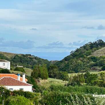 Uma paisagem rural tranquila com uma casa branca com telhado de telhas vermelhas, colinas verdejantes, árvores espalhadas e uma vista distante do céu azul.