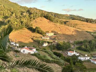 A luz dourada do sol banha um campo sereno com casas brancas espalhadas, colinas onduladas, árvores verdes e folhas de palmeira, criando uma cena rural pacífica ao pôr do sol.