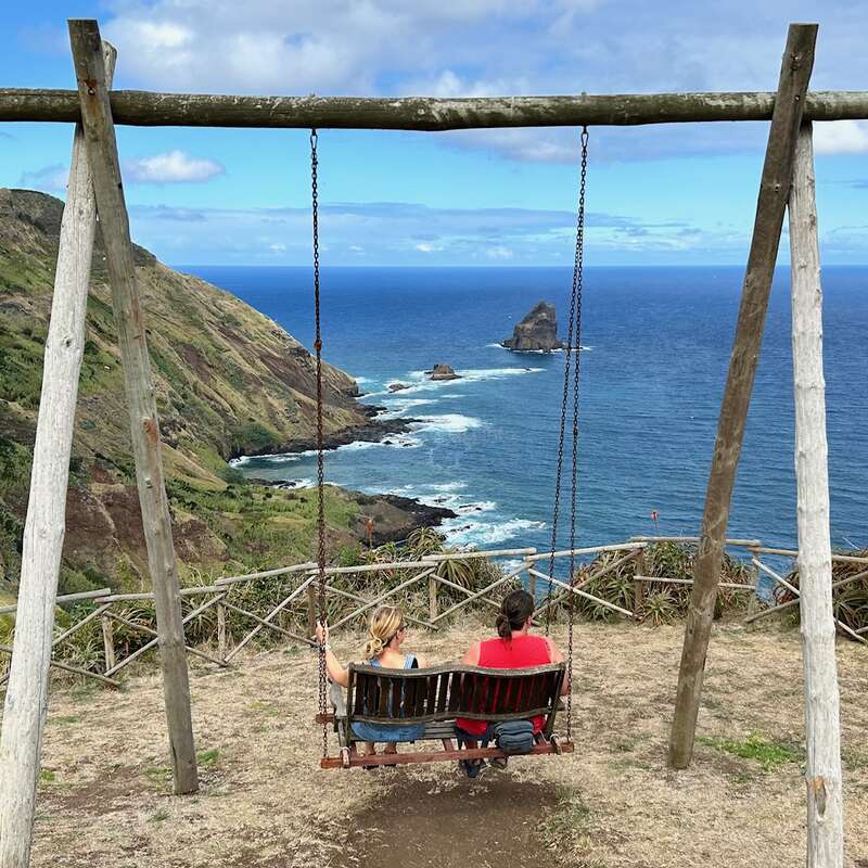 Duas pessoas sentam-se em um grande balanço de madeira com uma vista deslumbrante para o oceano, com penhascos escarpados, céu azul, rochas distantes e o mar que se estende infinitamente.