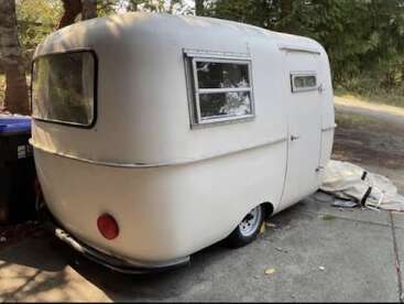 This image shows a small, vintage white camper trailer with rounded edges, parked on a driveway near trees. It has two windows and a rear red light.
