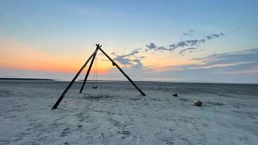 A minimalist beach scene at sunset shows a simple log swing structure on the sand, with soft colors in the sky and calm, tranquil atmosphere.
