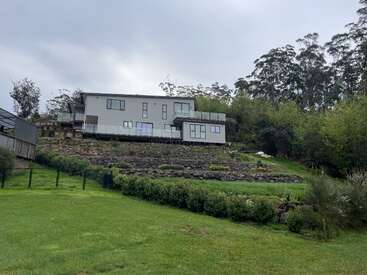 The image depicts a modern, two-story house situated on a hill, surrounded by trees and a lush green lawn, with a cloudy sky above.