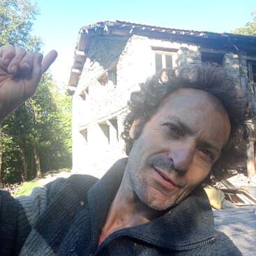 Ein Mann mit lockigem Haar macht ein Selfie im Freien und zeigt dabei nach oben. Er steht vor einem rustikalen Steinhaus, umgeben von Bäumen und hellem Sonnenlicht.