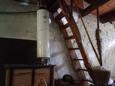 Ein gemütlicher, rustikaler Raum mit einer weißen Steinwand, einer Holztreppe, einem alten Wasserboiler, einem Fass, einigen Schränken und einem Paar nackter Füße.