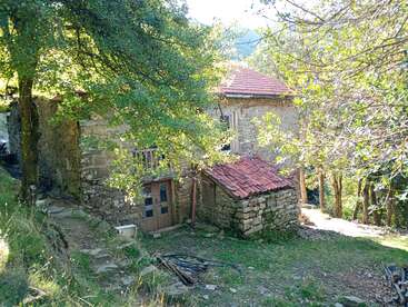 Ein altes Steinhaus mit roten Ziegeldächern steht in einem schattigen Waldgebiet. Das Sonnenlicht dringt durch die grünen Äste und wirft schummriges Licht auf das rustikale Gebäude.