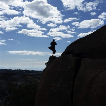 A imagem retrata uma cena serena de uma pessoa em uma pose de ioga, em pé sobre uma rocha, com os braços estendidos, tendo como pano de fundo um corpo de água e um céu claro.