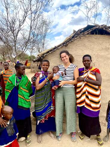 L'image représente une femme tenant un bébé, entourée d'un groupe de femmes et d'enfants portant des vêtements traditionnels, devant une hutte en terre dans un environnement rural.
