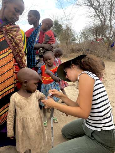Une femme portant une chemise rayée et un chapeau mesure la taille d'un enfant à l'aide d'un mètre ruban, entourée d'autres enfants et d'adultes dans une zone rurale.