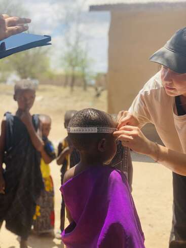 L'image représente une femme mesurant la tête d'un jeune enfant dans un village, avec d'autres enfants et un bâtiment visible à l'arrière-plan.