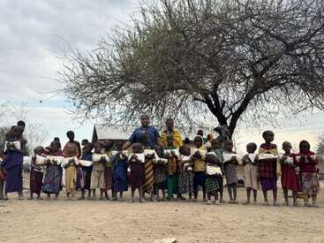 A group of children and adults stand outdoors holding packages, beneath a large tree. They appear happy and united, possibly during a community or aid event.