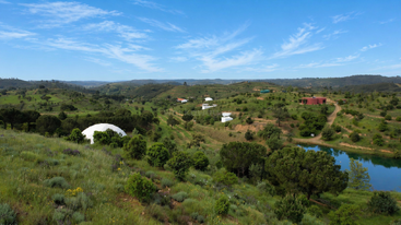 Des collines verdoyantes et vallonnées, parsemées d'arbres et d'arbustes, entourent des structures blanches en forme de dôme disséminées ici et là. Un étang reflète le ciel bleu, tandis que les collines lointaines s'étendent sous des nuages vaporeux.