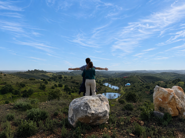 Deux personnes se tiennent au sommet d'une colline, les bras tendus, admirant le paysage. Une rivière serpente à travers les collines verdoyantes sous un vaste ciel bleu limpide.