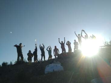 Un groupe de personnes se tient sur une colline, se découpant sur fond de soleil éclatant, les bras levés dans une gesticulation joyeuse, avec un ciel dégagé et de la végétation au premier plan.