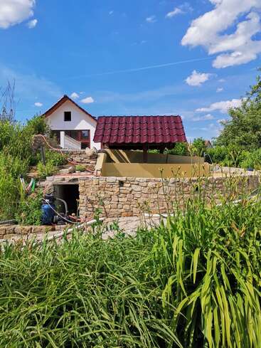 Eine malerische ländliche Szene mit weißem Haus, rotem Brunnen, Steinmauern, üppigen grünen Pflanzen, blauem Himmel und flauschigen weißen Wolken an einem sonnigen Tag.