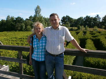 Ein lächelndes Paar steht auf einer Holzbrücke, umgeben von einem grünen Heckenlabyrinth und Bäumen, und genießt einen sonnigen Tag in einer schönen Umgebung.