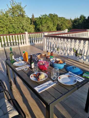 Ein schöner Esstisch im Freien ist für eine Mahlzeit mit Speisen, Getränken und Tellern gedeckt. Umgeben von Grün und einem eleganten weißen Geländer auf einer Terrasse.