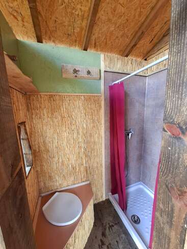 This image shows a rustic bathroom with a wooden ceiling, composting toilet, bamboo wall decor, small mirror, and a shower area with a red curtain. Cozy atmosphere.