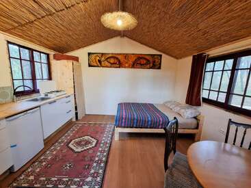 This cozy cabin features a wooden ceiling, bright kitchen, colorful rug, single bed with striped blanket, rustic table, large windows, and animal artwork above the bed.