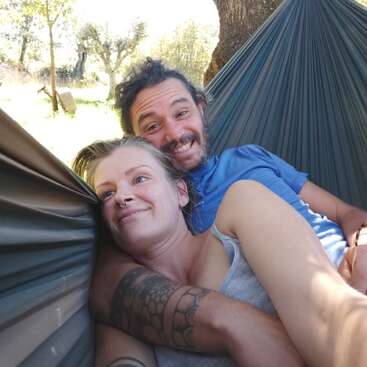 A happy couple relaxes together in a hammock outdoors. They are smiling, cuddling, and enjoying a sunny day with trees and nature in the background.