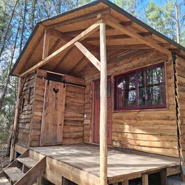 A small rustic wooden cabin stands in a forest, featuring a porch, red-framed windows, wooden stairs, and a charming door with a heart-shaped cutout.