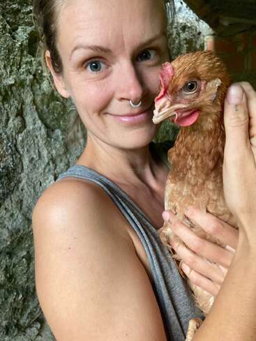 Une femme avec un anneau dans le nez sourit chaleureusement en tenant un poulet brun. Elle porte un débardeur gris et ils semblent proches et à l'aise ensemble en plein air.