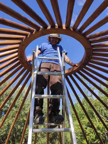 Un homme se tient sur une échelle à l'intérieur d'une charpente circulaire en bois, sous un ciel bleu, souriant, entouré d'arbres et de soleil, en train de construire une yourte.