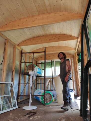 Un homme marche à l'intérieur d'une pièce partiellement construite avec un plafond en bois incurvé. Des matériaux de construction, des outils, un ventilateur et un cadre de fenêtre sont éparpillés sur le sol.
